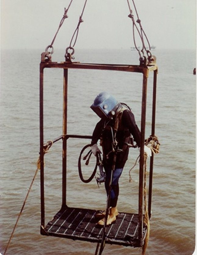 Diver in cage suspended over water.
